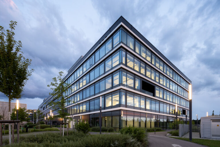 Modern glass-fronted office building at dusk with illuminated interiors and surrounding landscaping in the foreground.