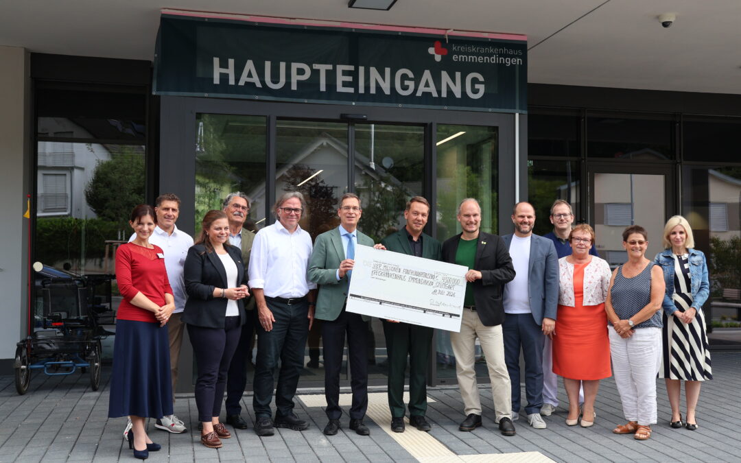 Group of professionals standing outside the hospital main entrance under a 'HAUPTEINGANG' sign, holding a large ceremonial check.