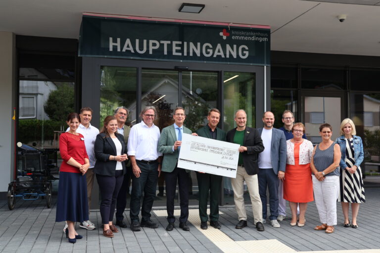 Group of professionals standing outside the hospital main entrance under a 'HAUPTEINGANG' sign, holding a large ceremonial check.