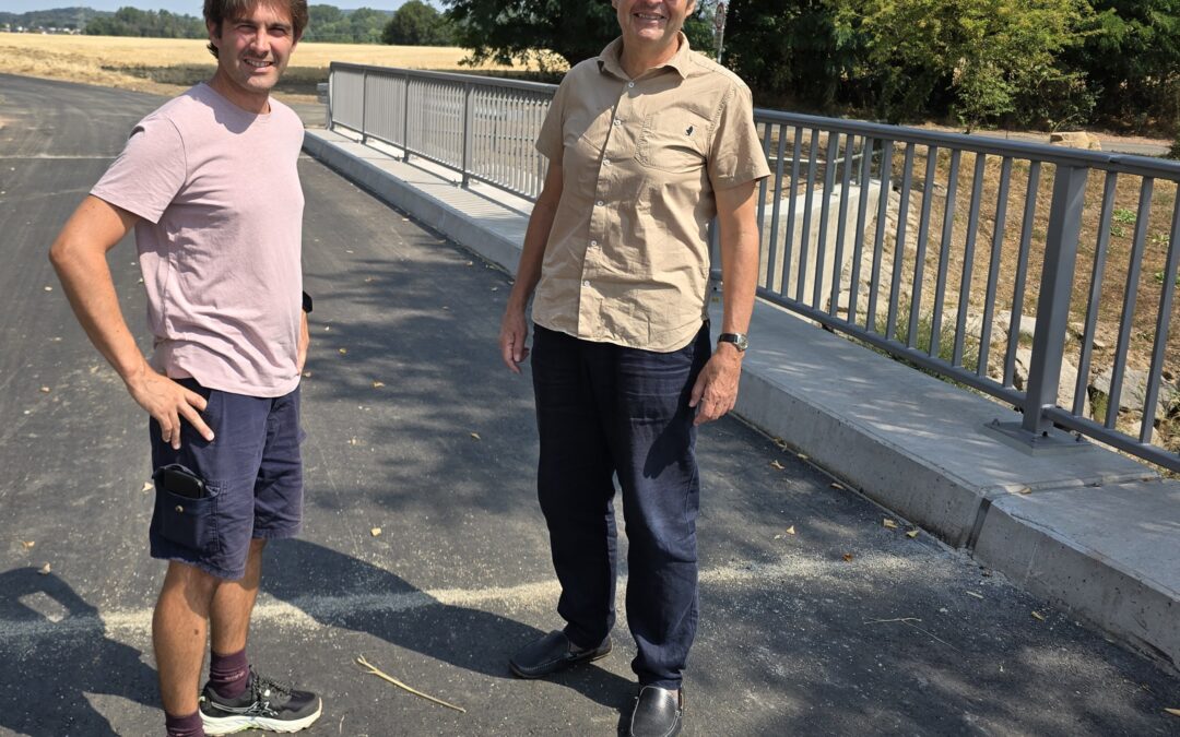 Two men stand outdoors on a sunny day beside a metal railing, smiling, with a rural field behind them.