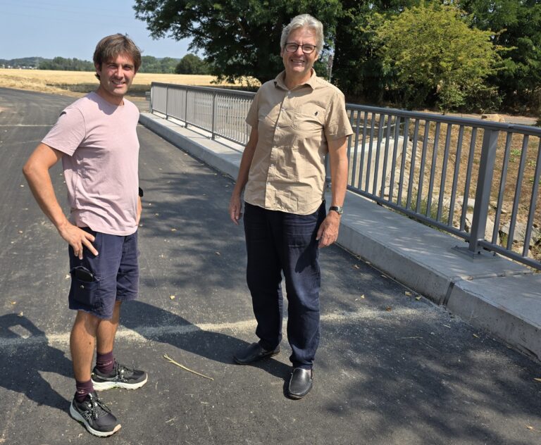 Two men stand outdoors on a sunny day beside a metal railing, smiling, with a rural field behind them.
