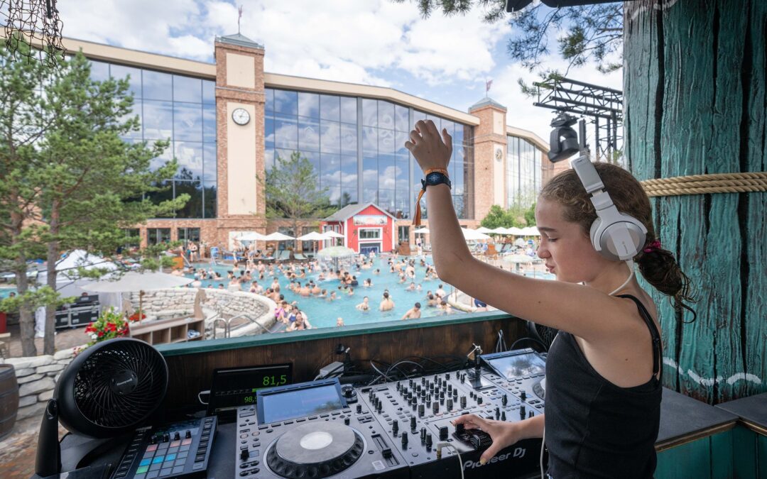 DJ wearing headphones, operating a Pioneer DJ mixer at a lively poolside party with a cheering crowd in the background. (Informative)