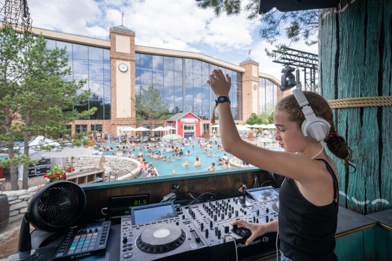DJ wearing headphones, operating a Pioneer DJ mixer at a lively poolside party with a cheering crowd in the background. (Informative)