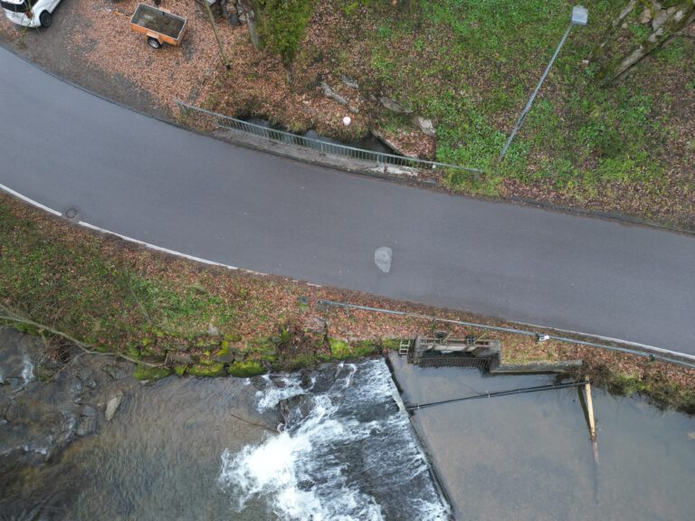 Aerial view of a river with a small waterfall beside a curved road and metal railing, surrounded by fallen leaves and green vegetation.