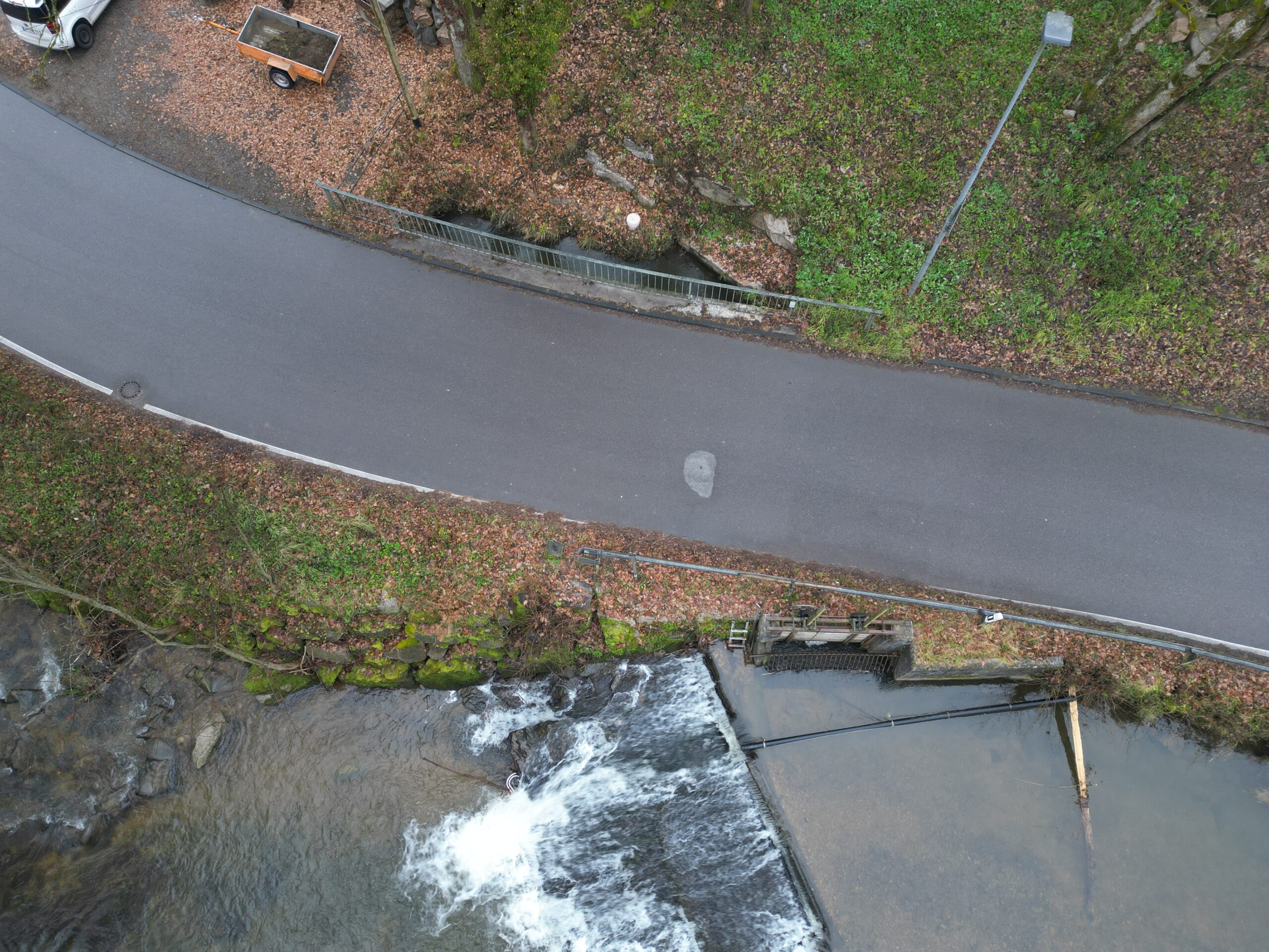 Aerial view of a river with a small waterfall beside a curved road and metal railing, surrounded by fallen leaves and green vegetation.