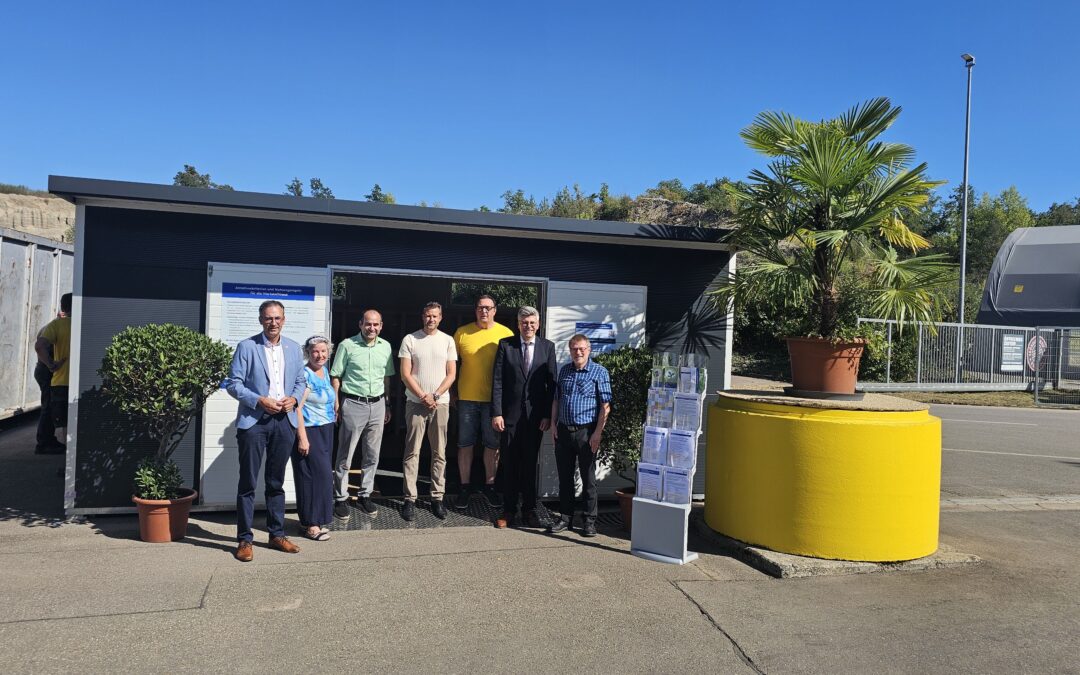 Eight adults stand together outside a small modern building with signs, posing for a group photo on a sunny day.