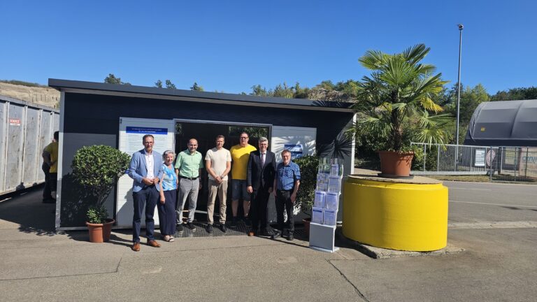 Eight adults stand together outside a small modern building with signs, posing for a group photo on a sunny day.
