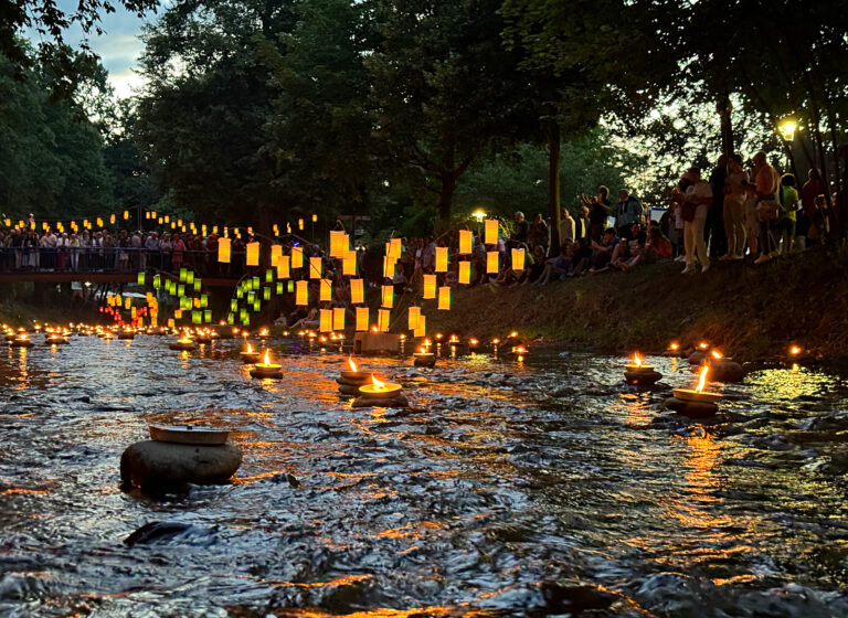 Crowd gathered along a riverbank at dusk while glowing orange lanterns float on the water and float-lit decorations illuminate the scene.