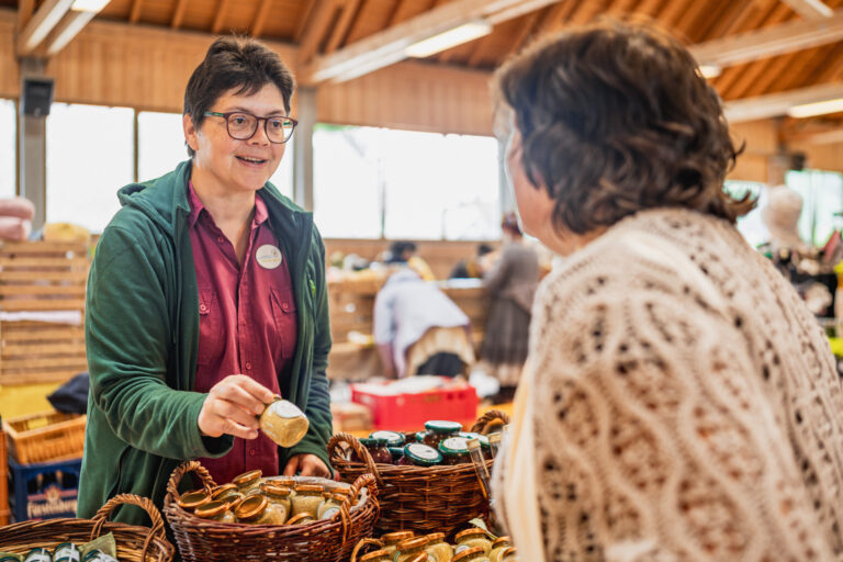 Vendor with short dark hair and glasses talks to a customer at a market stall filled with jars and woven baskets.