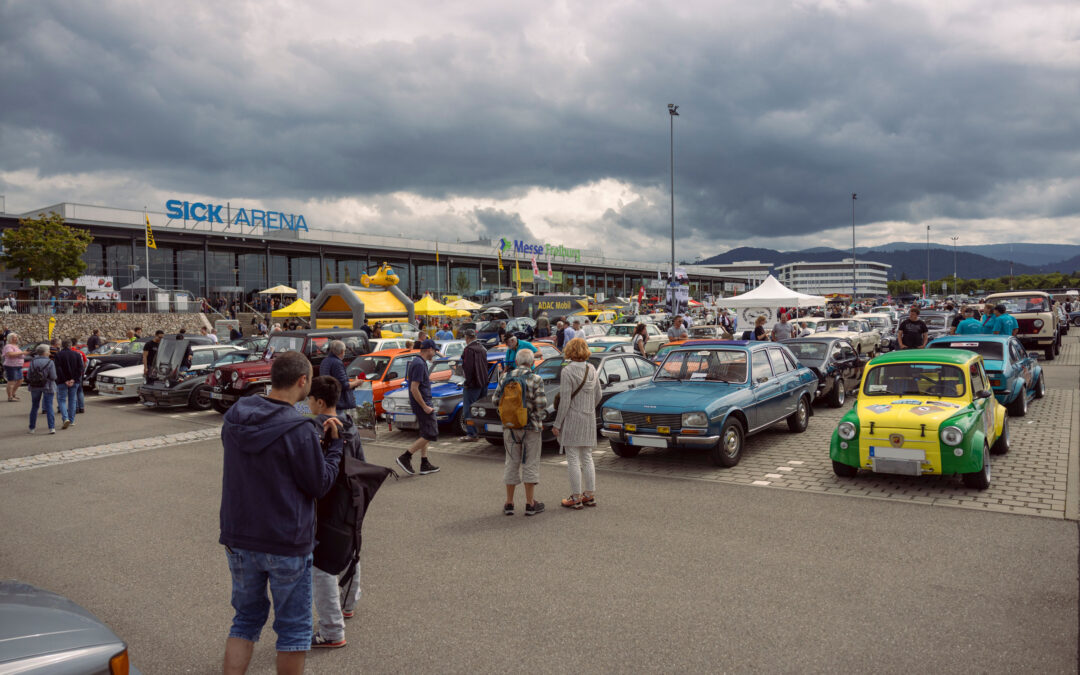 Car show crowd outside a large venue with vintage cars lined up and tents in the foreground under a cloudy sky