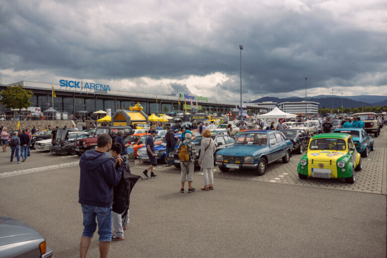 Car show crowd outside a large venue with vintage cars lined up and tents in the foreground under a cloudy sky