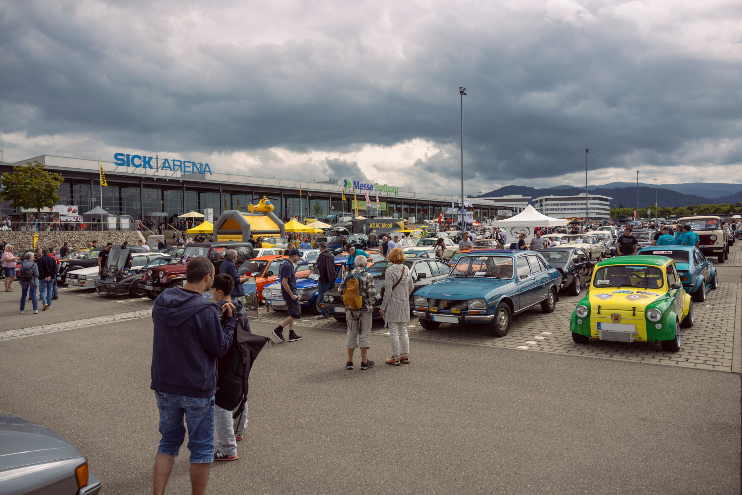 Car show crowd outside a large venue with vintage cars lined up and tents in the foreground under a cloudy sky