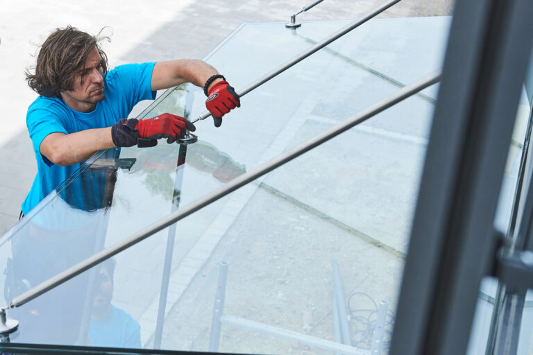 Man in a bright blue shirt wearing red gloves tightens a clamp on a glass railing outdoors.