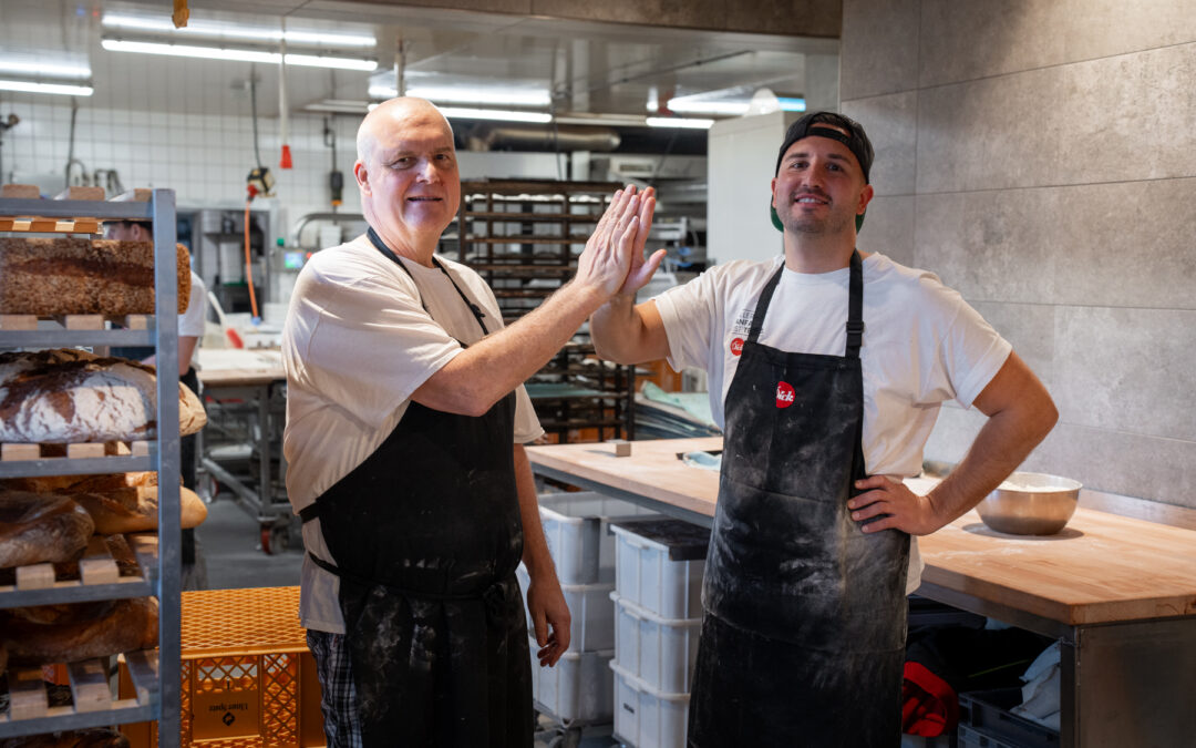 Two bakers in aprons share a high-five in a busy bakery kitchen with loaves on racks nearby.