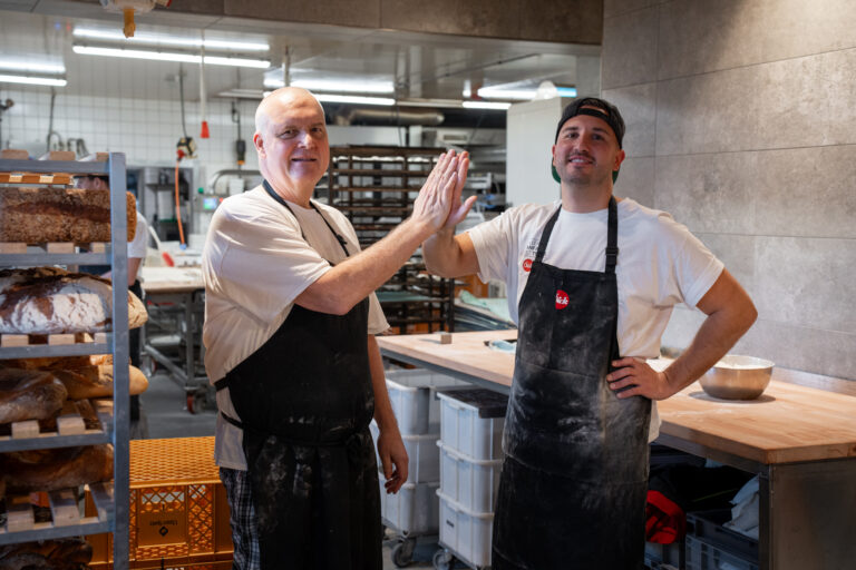 Two bakers in aprons share a high-five in a busy bakery kitchen with loaves on racks nearby.