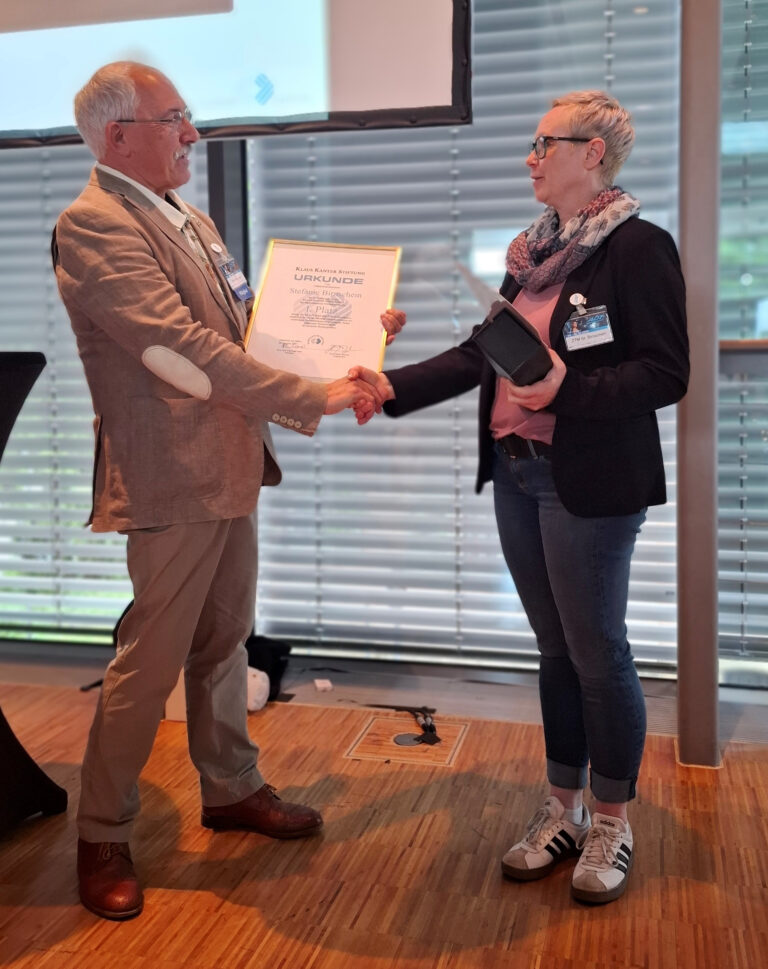 Two people in a formal setting shake hands; a man presents a framed certificate to a woman who holds a tablet or folder, indoors by blinds with a wooden floor.