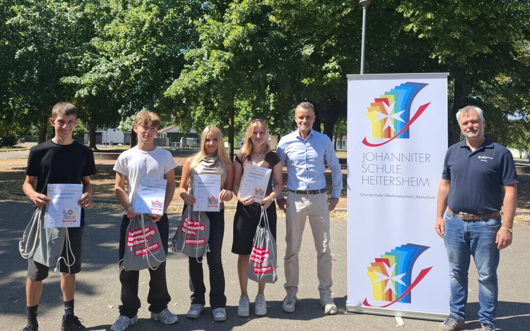 Group of six people standing outdoors, four young students holding certificates and bags, with two adults and a tall banner for Johanniter Schule Heitersheim nearby.