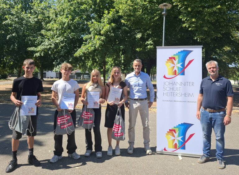 Group of six people standing outdoors, four young students holding certificates and bags, with two adults and a tall banner for Johanniter Schule Heitersheim nearby.