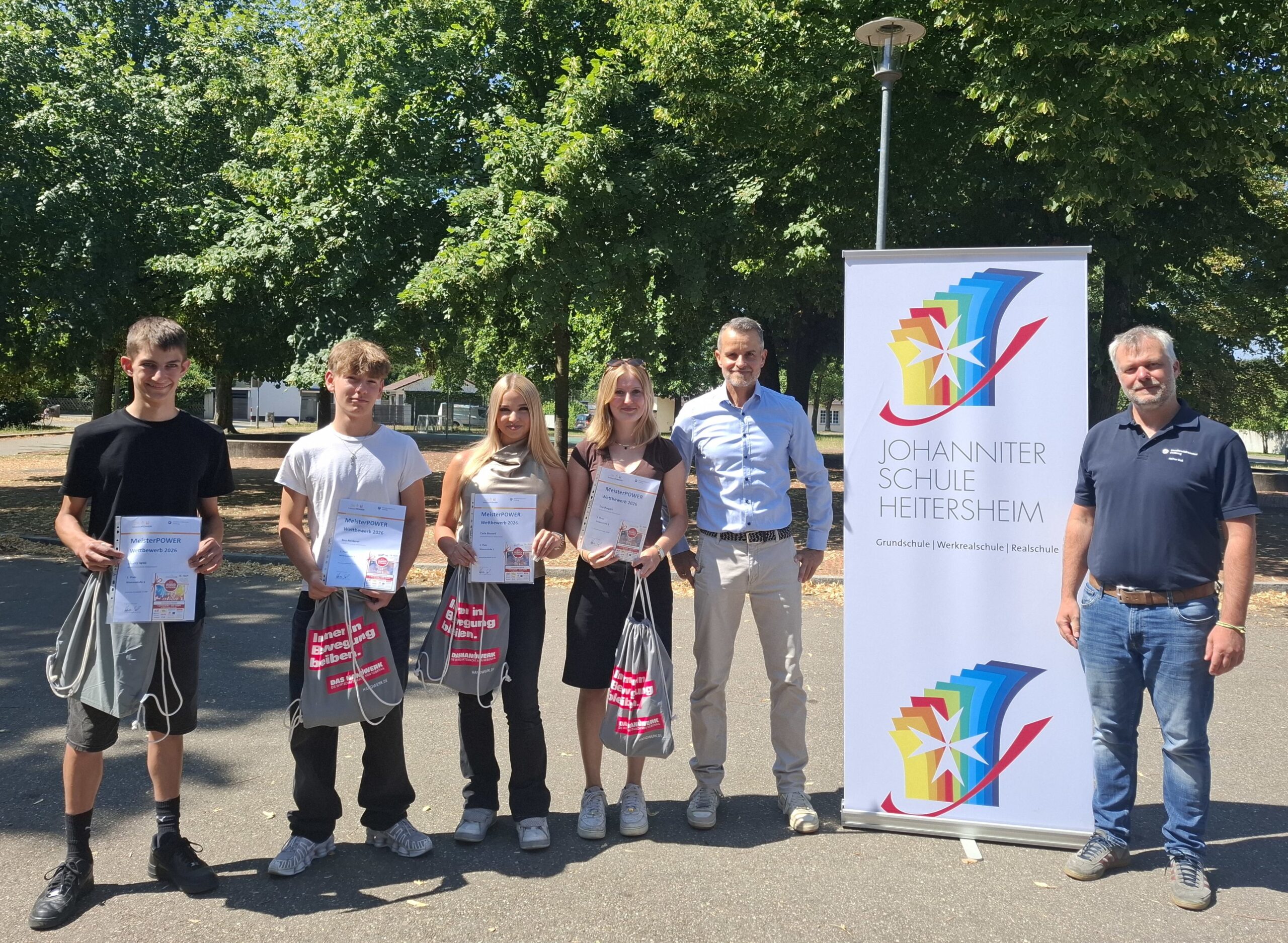Group of six people standing outdoors, four young students holding certificates and bags, with two adults and a tall banner for Johanniter Schule Heitersheim nearby.