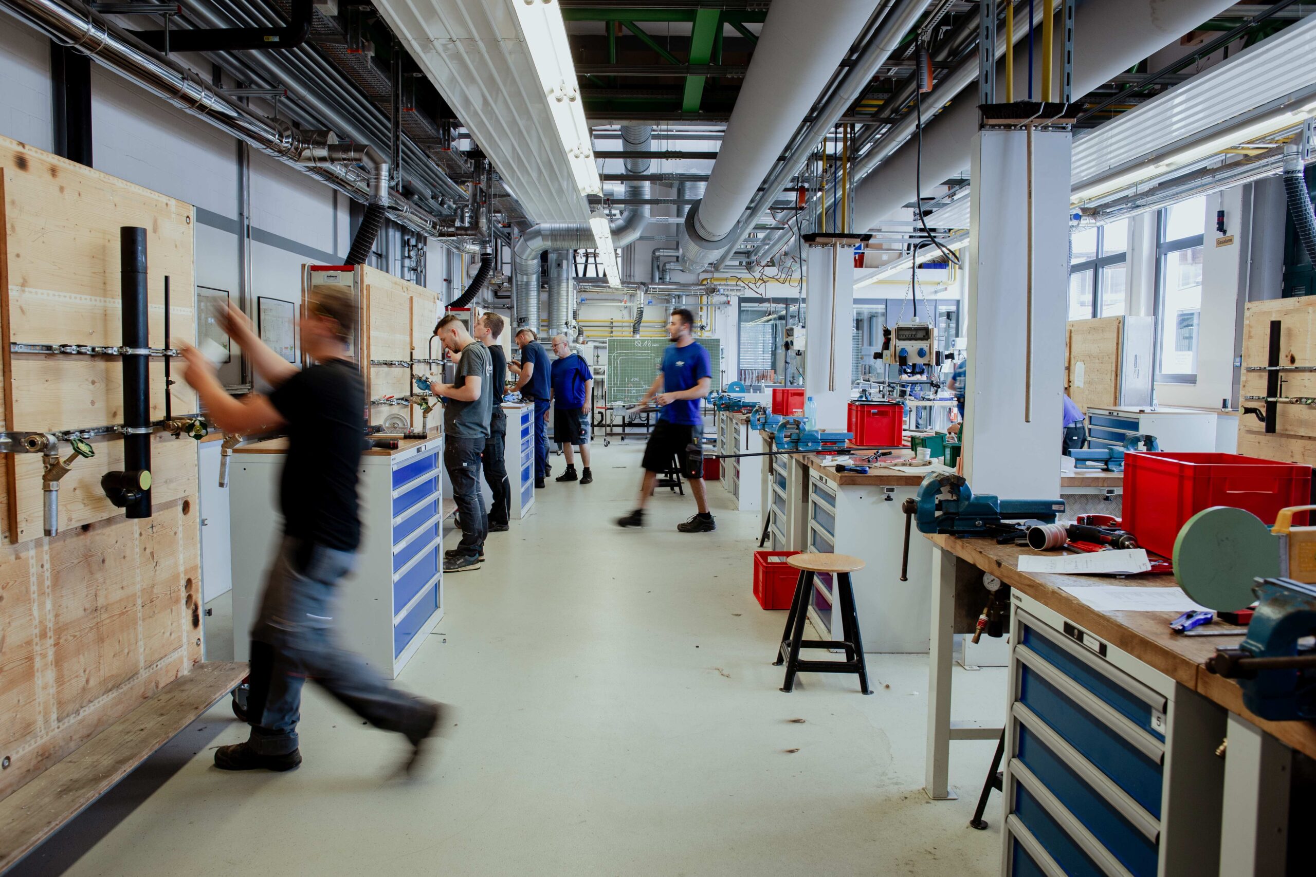 Several technicians work at benchtop stations in a well-lit industrial workshop filled with tools and equipment. (informative)