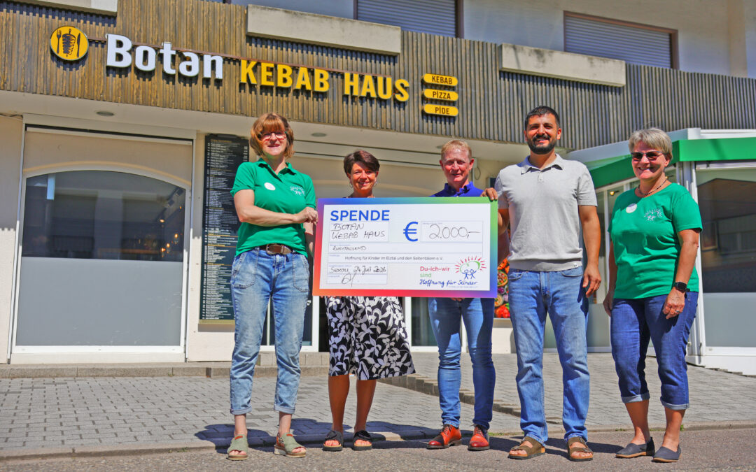 Five adults stand outside a kebab shop, holding a large donation check and smiling at the camera.