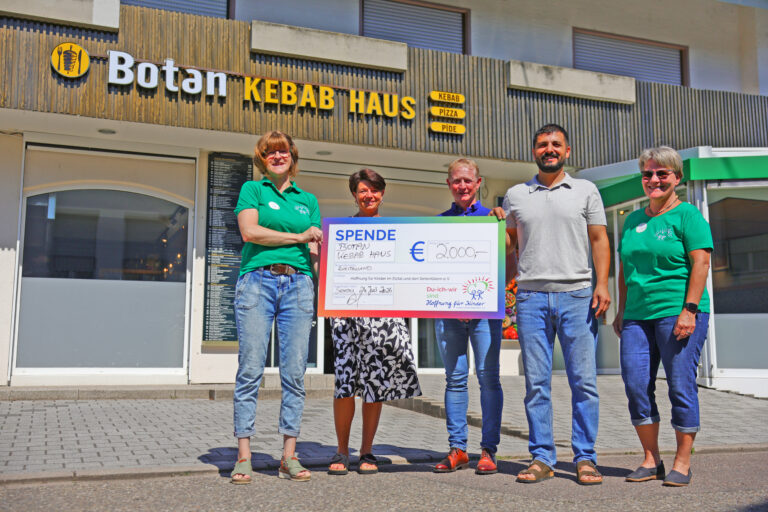 Five adults stand outside a kebab shop, holding a large donation check and smiling at the camera.