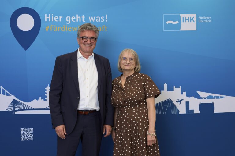 Two adults posing in front of a large blue event backdrop: a man in a dark suit and white shirt beside a woman in a brown polka-dot dress, both smiling.