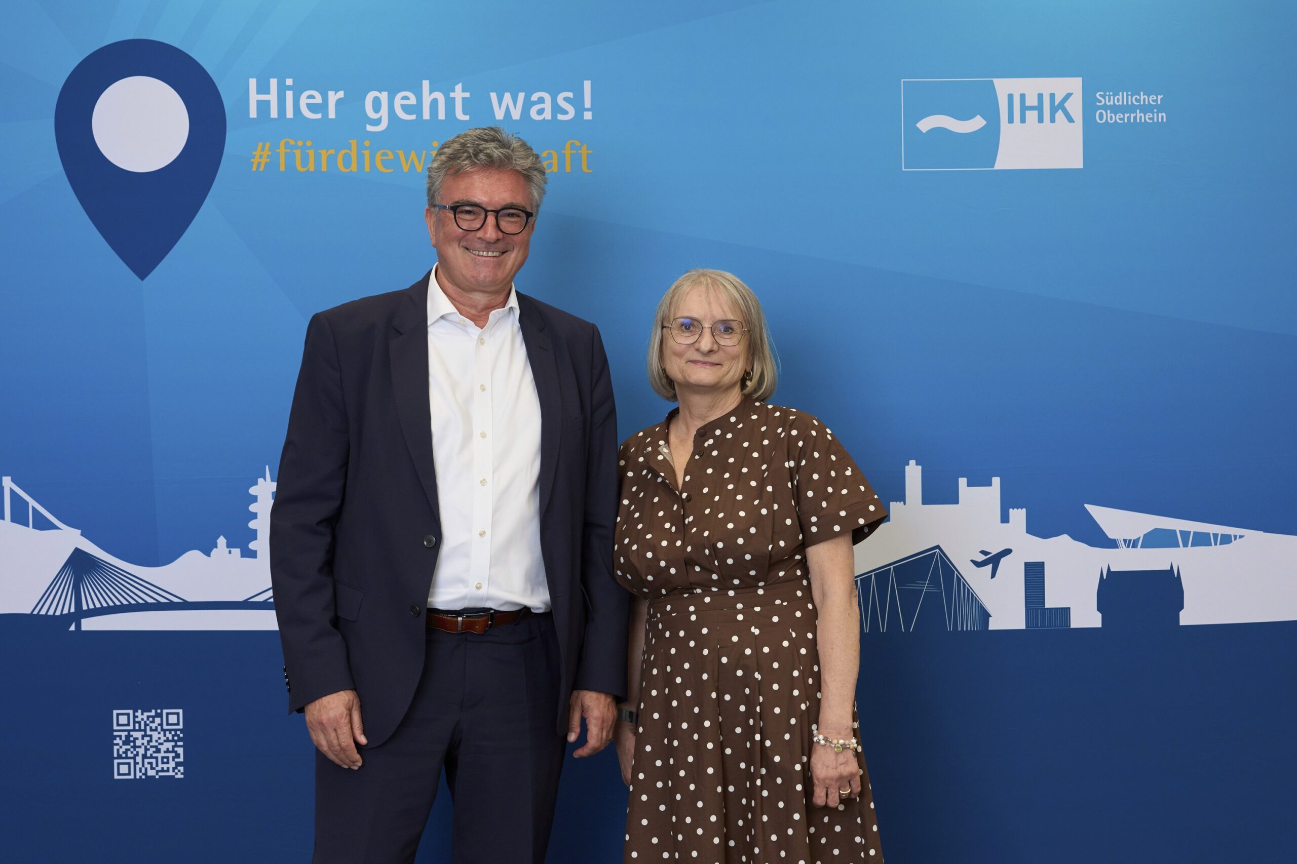 Two adults posing in front of a large blue event backdrop: a man in a dark suit and white shirt beside a woman in a brown polka-dot dress, both smiling.