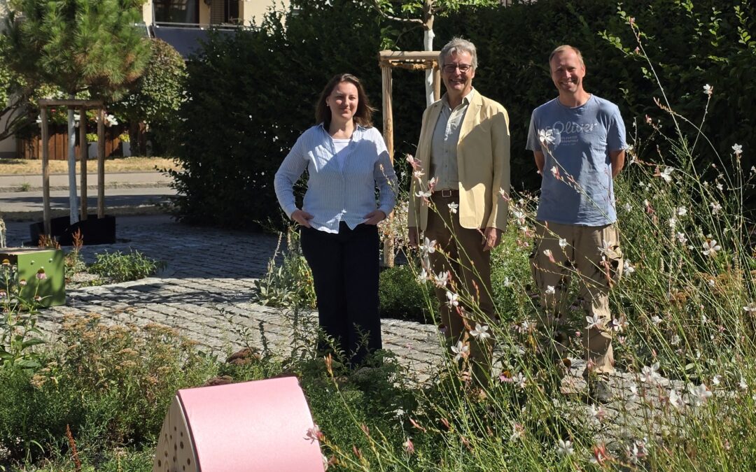 Three adults pose in a sunny garden with a pink curved wooden sculpture in the foreground, greenery and buildings in the background.