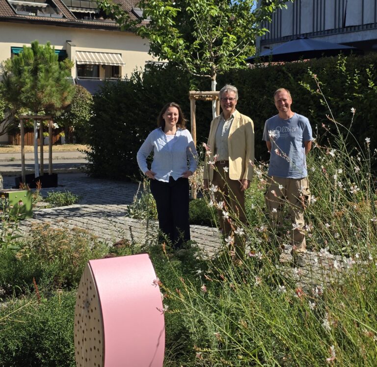 Three adults pose in a sunny garden with a pink curved wooden sculpture in the foreground, greenery and buildings in the background.