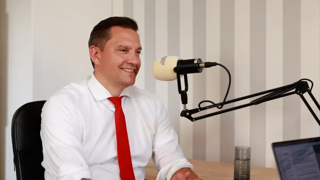 Man in a white shirt and red tie speaks into a professional podcast microphone in a studio setting, smiling.