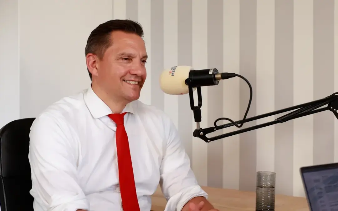 Man in a white shirt and red tie speaks into a professional podcast microphone in a studio setting, smiling.