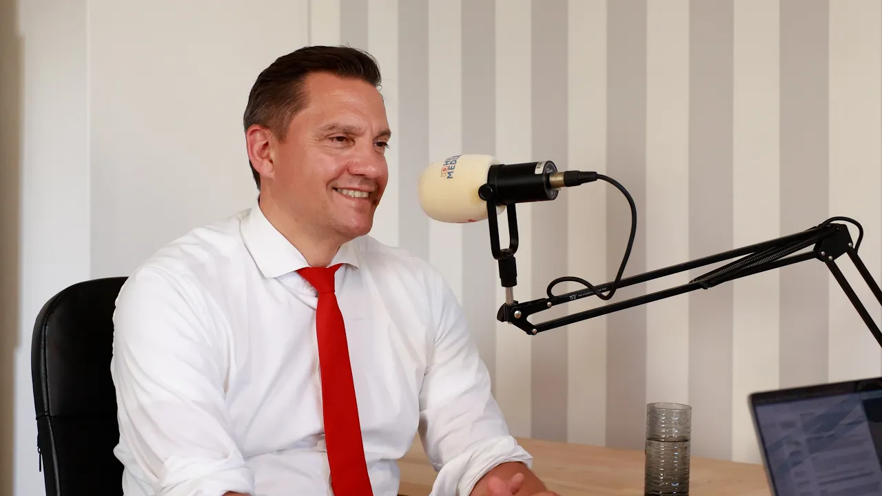 Man in a white shirt and red tie speaks into a professional podcast microphone in a studio setting, smiling.