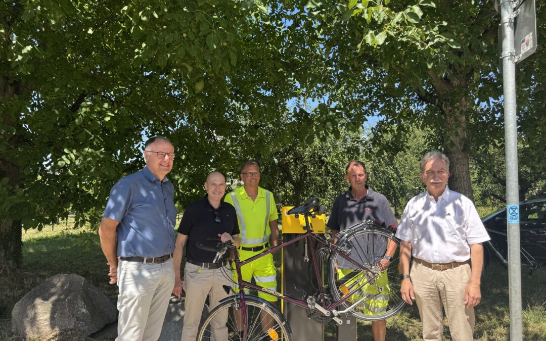 Group of six men posing outdoors under a leafy tree, with bicycles and a yellow bike rack nearby.