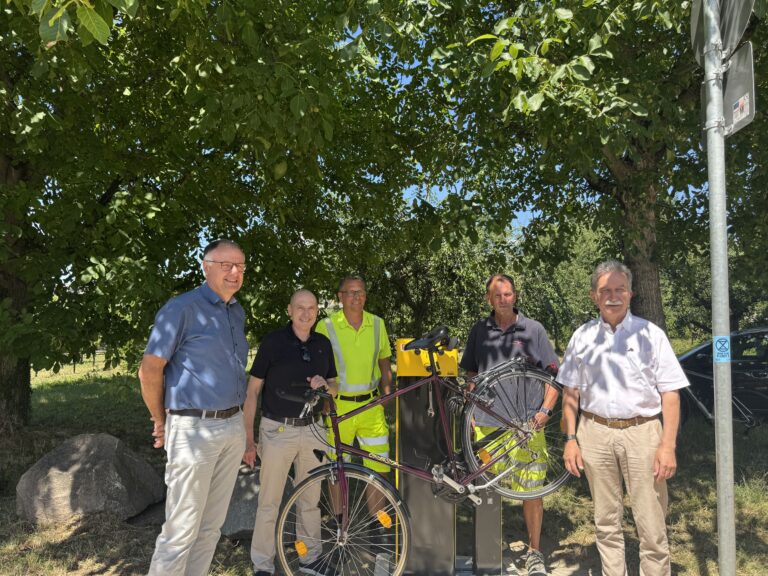 Group of six men posing outdoors under a leafy tree, with bicycles and a yellow bike rack nearby.