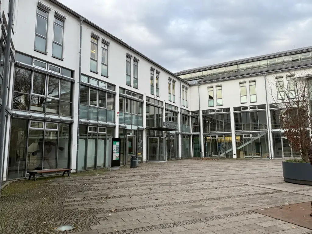 Modern white office building with large glass windows in a paved courtyard, under a cloudy sky.
