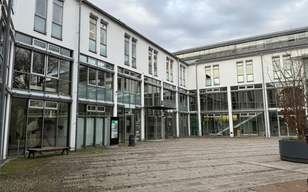 Modern white office building with large glass windows in a paved courtyard, under a cloudy sky.