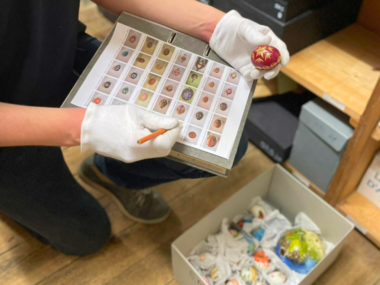 Person wearing white gloves holding a clipboard with a sheet showing many small decorative items, plus a box of crumpled paper nearby.