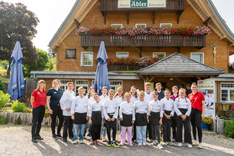 Group of people in white chef jackets posing outside a chalet-style hotel with flower-filled balconies and a sign reading Hotel Adler Bärenatal behind them.