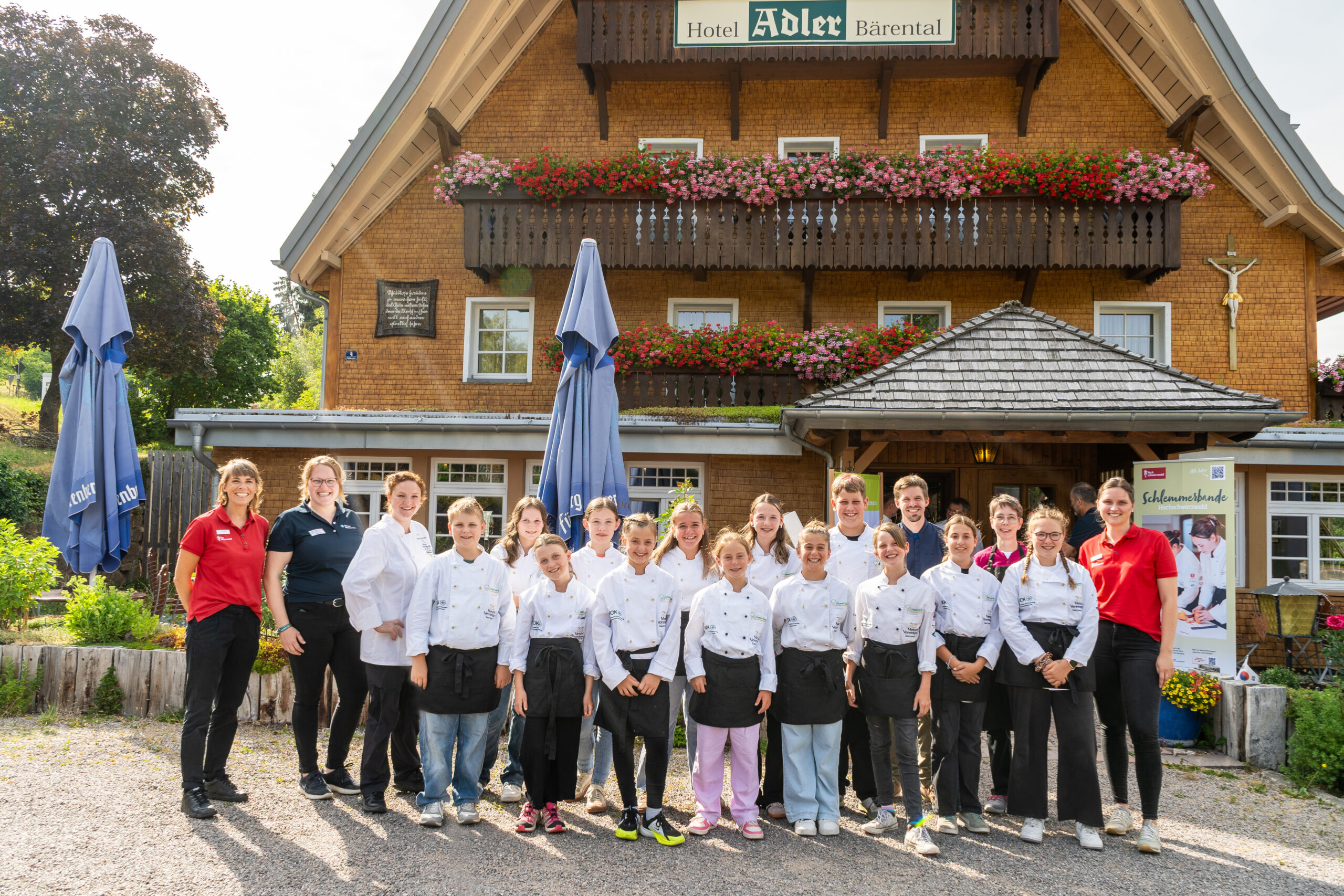 Group of people in white chef jackets posing outside a chalet-style hotel with flower-filled balconies and a sign reading Hotel Adler Bärenatal behind them.