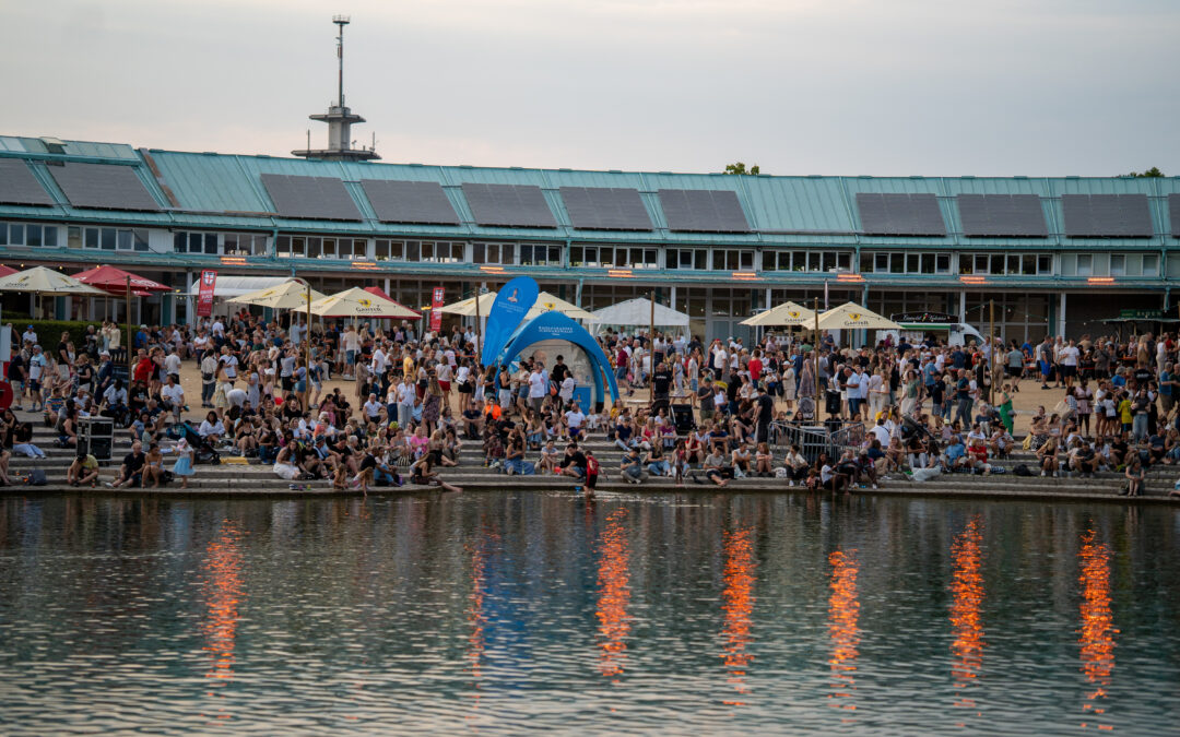 Crowd of people at a riverside festival with white umbrellas, seated on steps, beside a turquoise building with solar panels in the background