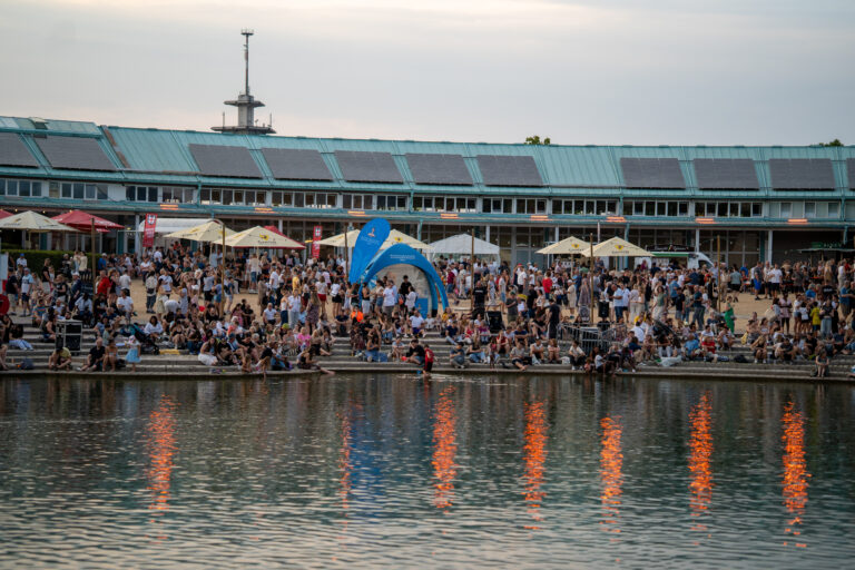 Crowd of people at a riverside festival with white umbrellas, seated on steps, beside a turquoise building with solar panels in the background