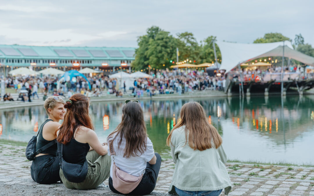 Four friends sit on a stone waterfront, watching a crowded outdoor festival with tents and string lights in the background.