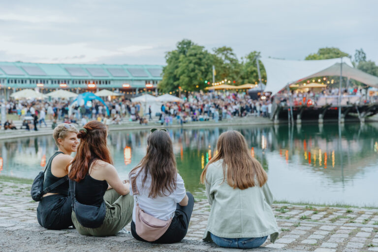 Four friends sit on a stone waterfront, watching a crowded outdoor festival with tents and string lights in the background.