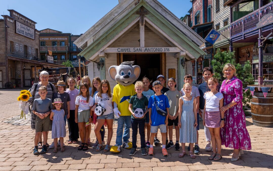 Group of children posing with a large mouse mascot in a rustic Old West town square, smiling at the camera.