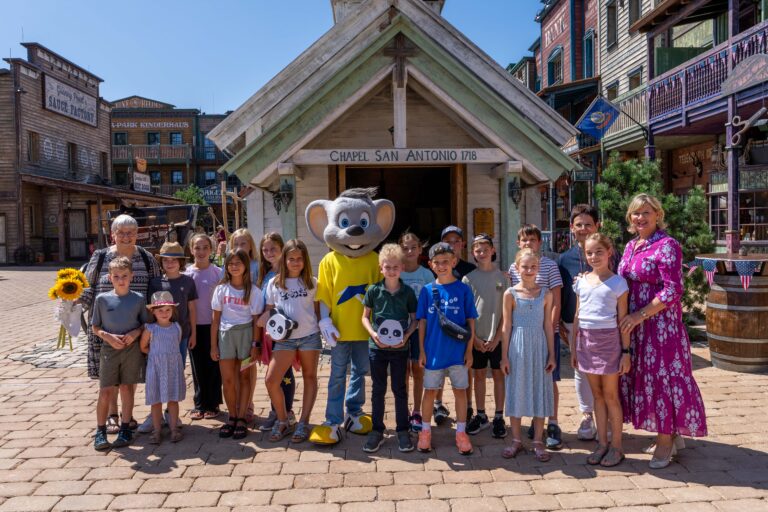 Group of children posing with a large mouse mascot in a rustic Old West town square, smiling at the camera.