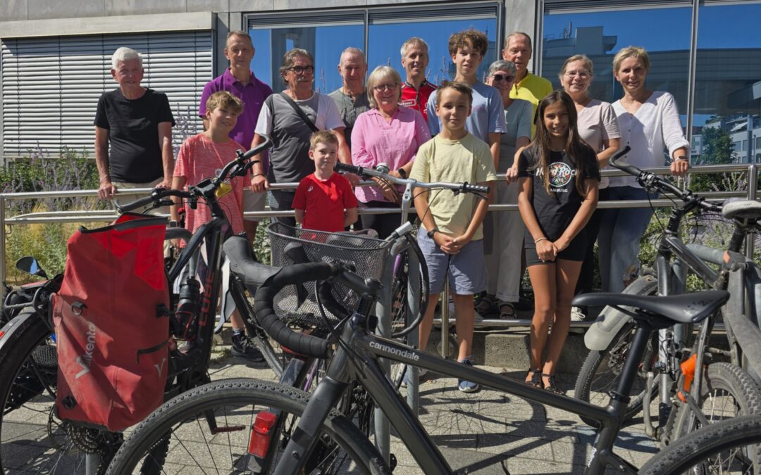 Group of people of varying ages standing behind parked bicycles outside a modern glass building.