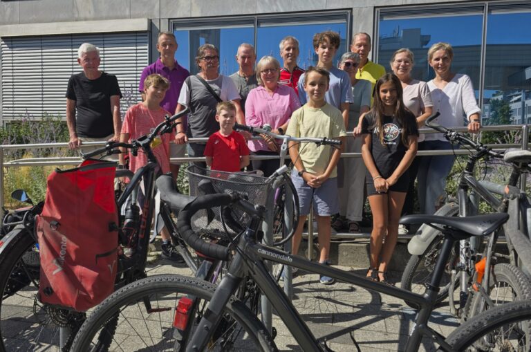 Group of people of varying ages standing behind parked bicycles outside a modern glass building.