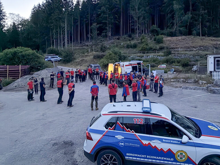 Group of emergency responders in red vests forming a circle for briefing in a parking area, with an ambulance and forested hillside in the background.