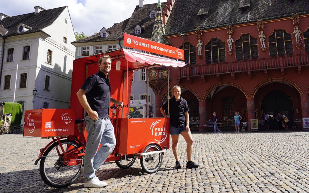 Two people stand beside a bright red tour information bike cart in a cobblestone square, with historic buildings in the background.